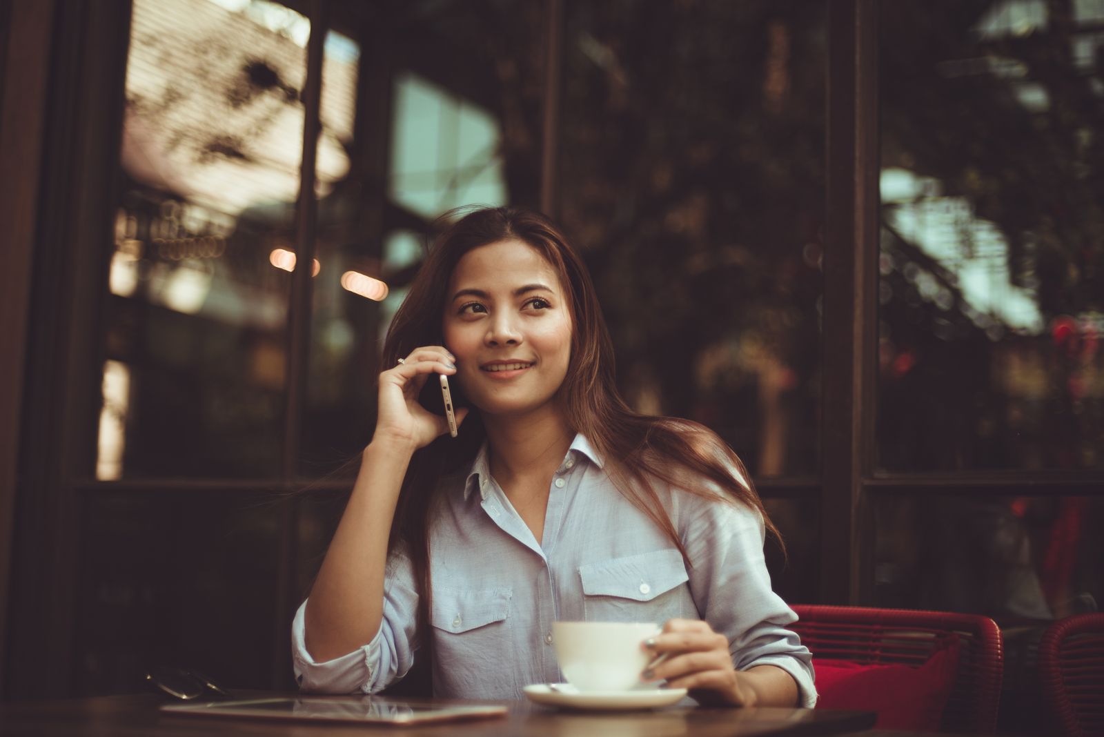 Mulher sorridente de camisa azul falando ao telefone em uma mesa de café, com uma xícara ao lado.